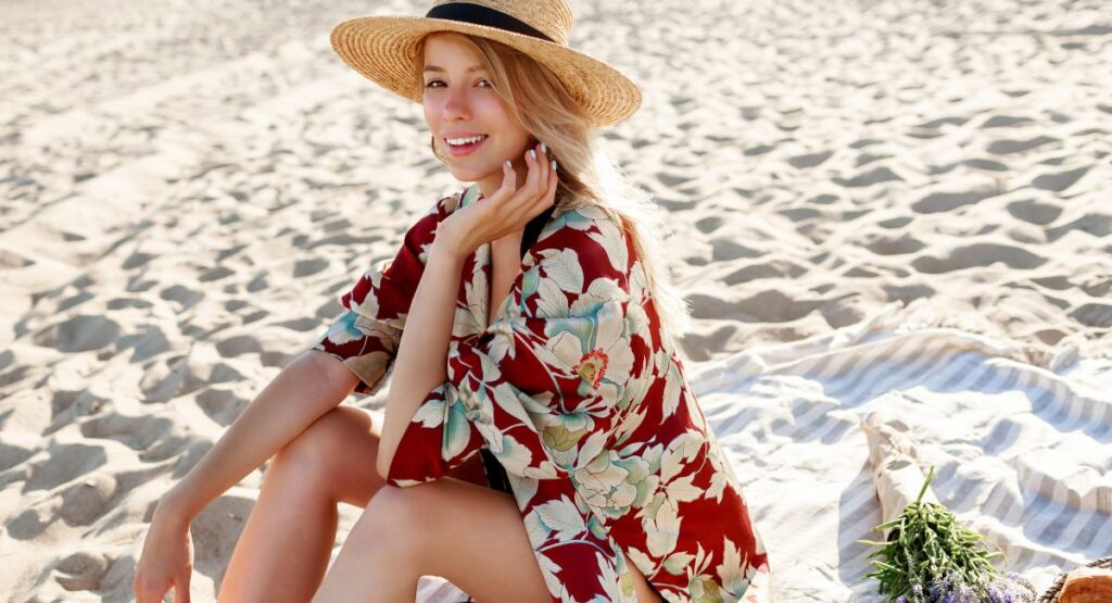 Woman in red floral dress sitting on sandy beach with straw hat demonstrating complete sun protection with Sunbetter sunscreen