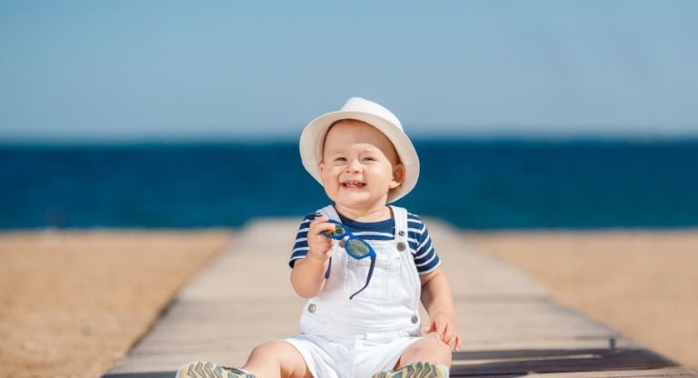 Baby wearing essential newborn sun protection clothing with a white sun hat and striped shirt at the beach, demonstrating proper UV safety for infants.