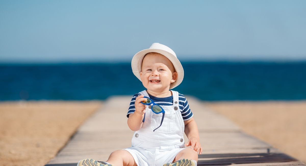 Baby wearing essential newborn sun protection clothing with a white sun hat and striped shirt at the beach, demonstrating proper UV safety for infants.