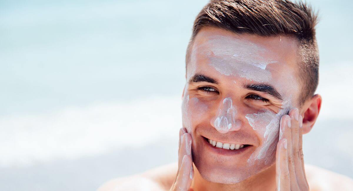 Alt text: "How often to reapply sunscreen on face - smiling man applying white mineral sunscreen properly on facial areas most prone to sun damage