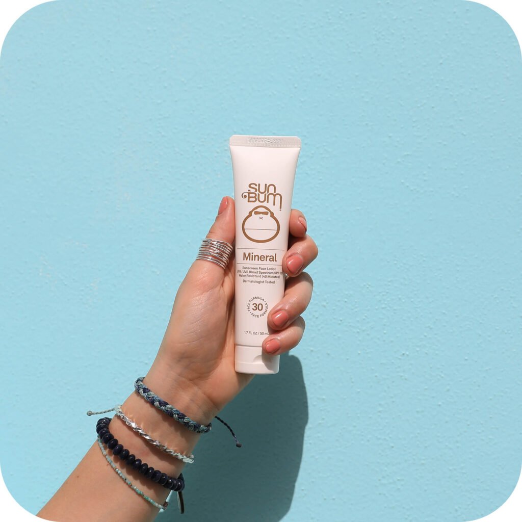 Hand holding Sun Bum Mineral SPF 30 sunscreen tube against turquoise background, showing if Sun Bum sunscreen is good for sensitive skin