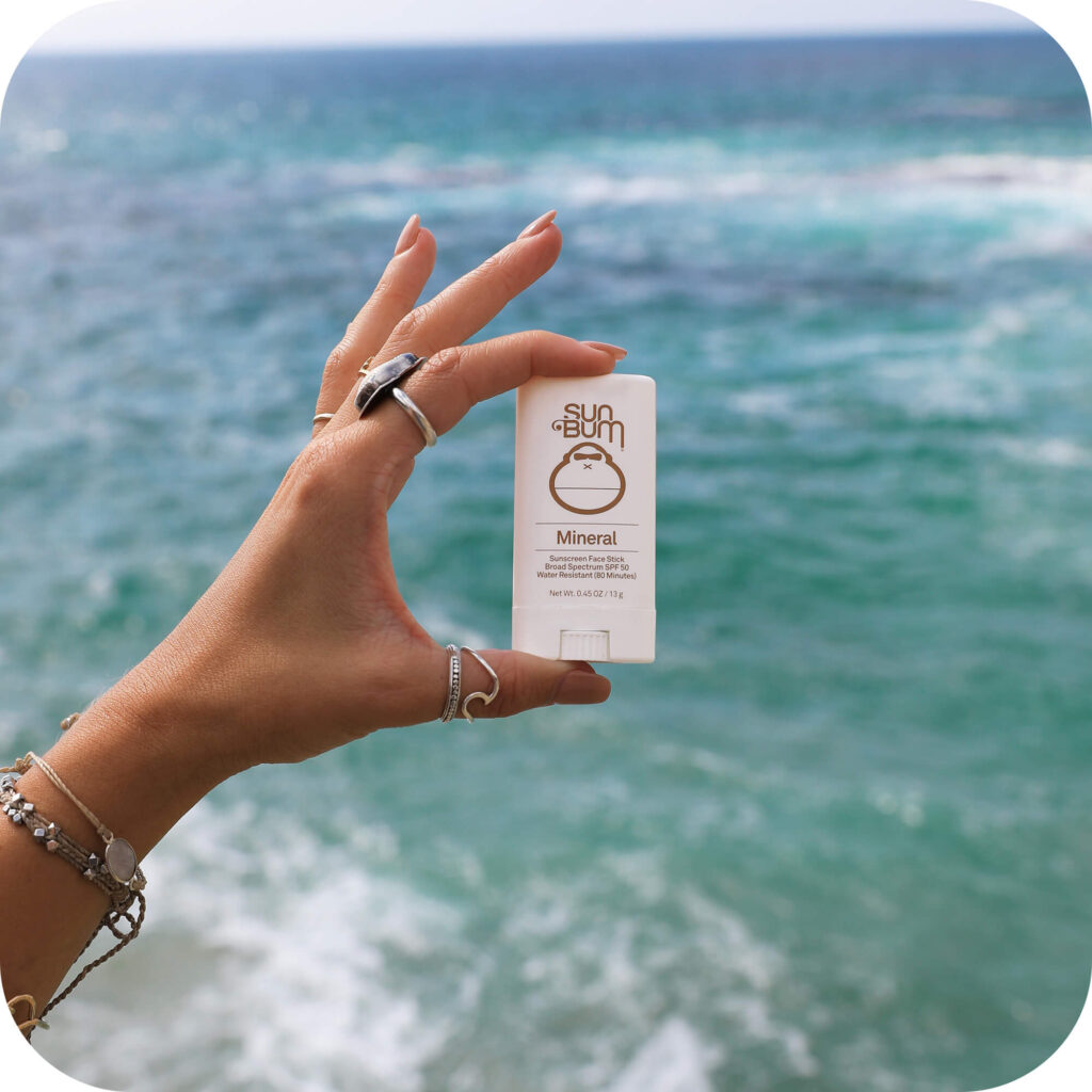 Sun Bum Mineral Face Stick SPF 50 held up against ocean backdrop, perfect for on-the-go protection at the beach