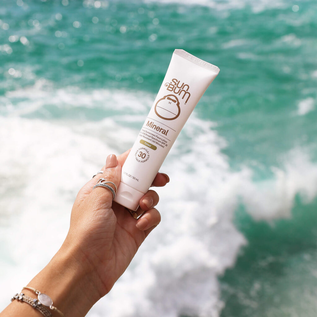 Person holding Sun Bum Mineral sunscreen with SPF 30 by the ocean, demonstrating why Sun Bum sunscreen is good for water activities