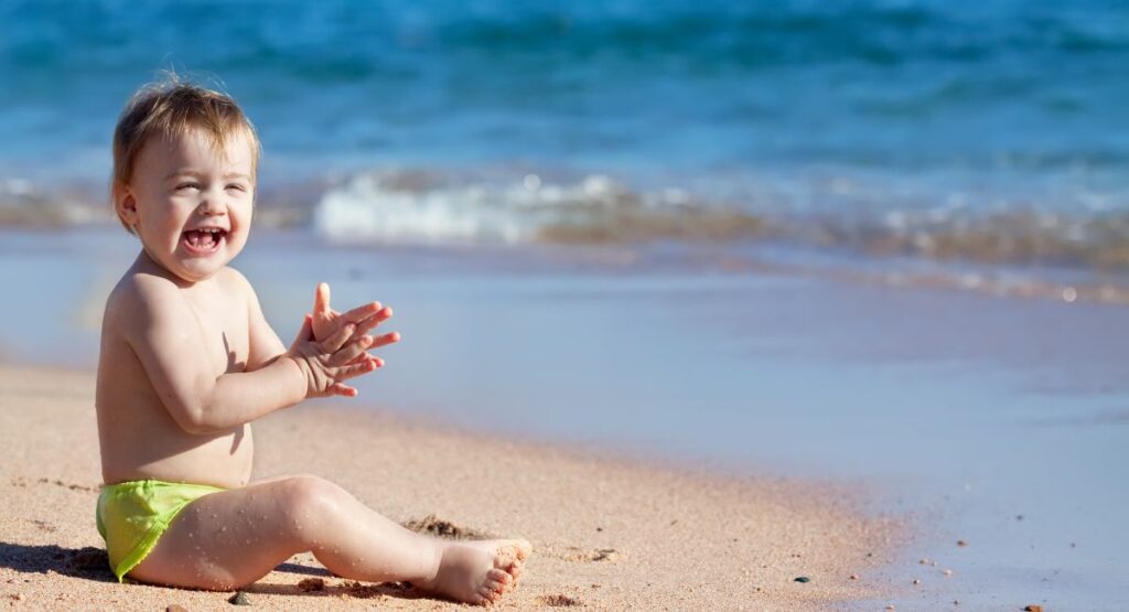 Happy toddler sitting on beach sand wearing bright lime green sun protection swim bottoms, clapping hands with ocean waves in the background.