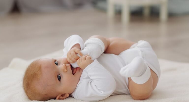Infant in all-white sun protective clothing including mittens lying on back on soft white blanket, exploring hands and feet.