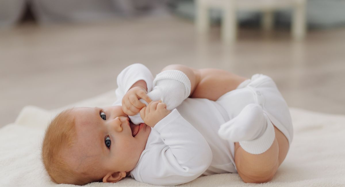Infant in all-white sun protective clothing including mittens lying on back on soft white blanket, exploring hands and feet.