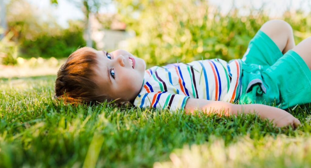 Young child with light brown hair relaxing on green grass wearing striped shirt and teal shorts, enjoying outdoor sunshine in summer.
