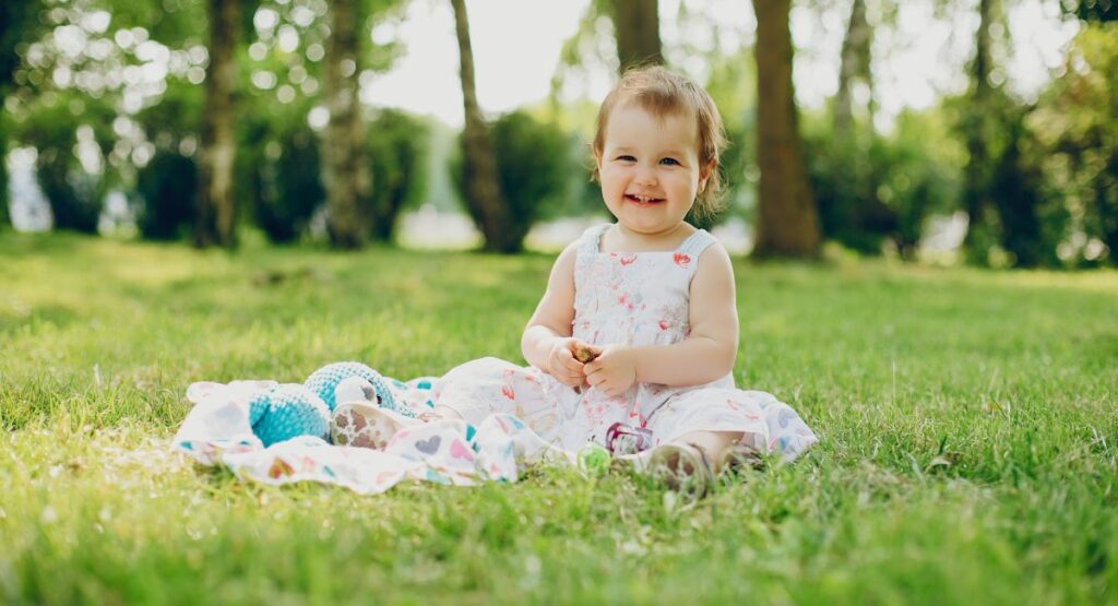 Toddler girl in white floral pattern sun dress sitting on grass with patterned blanket and toys, smiling in a park with trees in background.