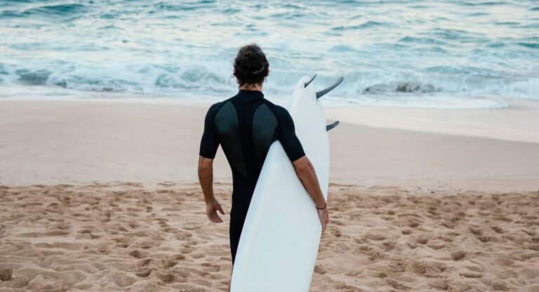 Person wearing a black short-sleeve wetsuit for sun protection standing on sandy beach, holding white surfboard while facing ocean waves - quality sun protection clothing swimwear for water sports enthusiasts.