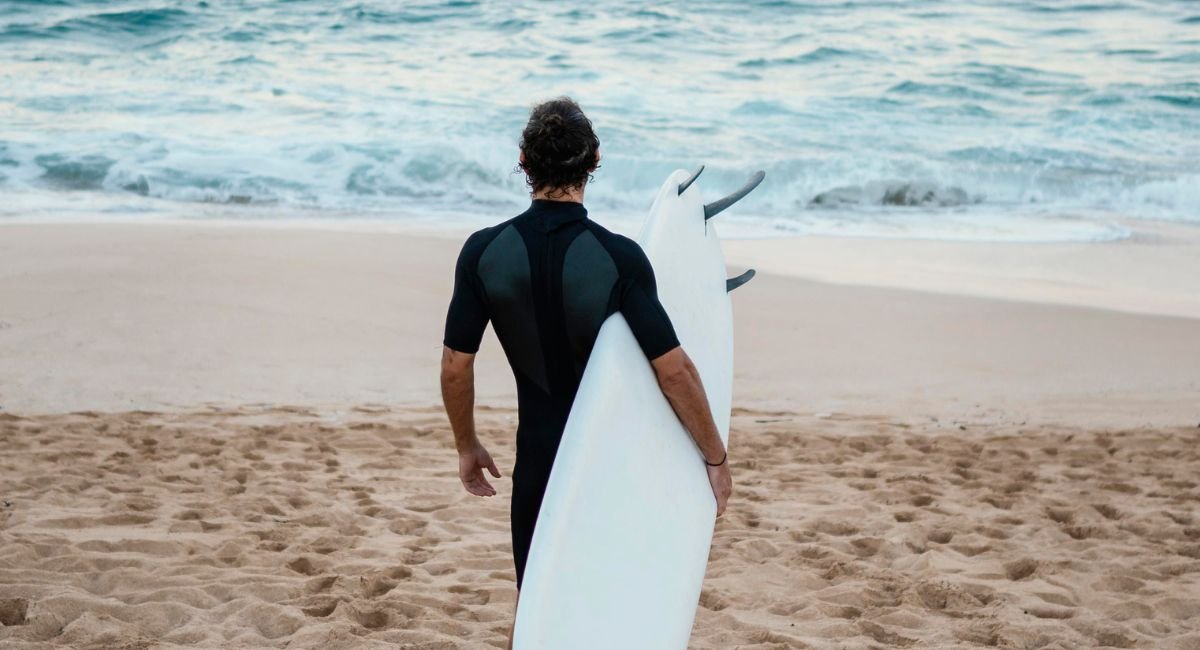 Person wearing a black short-sleeve wetsuit for sun protection standing on sandy beach, holding white surfboard while facing ocean waves - quality sun protection clothing swimwear for water sports enthusiasts.