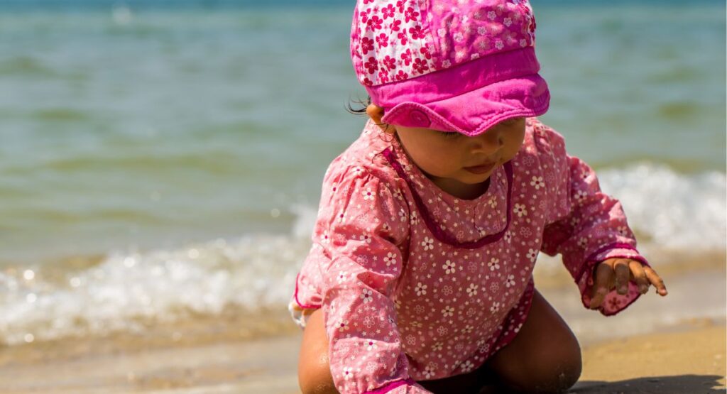 Baby wearing pink floral pattern sun protection clothing with matching sun hat crawling on beach sand with gentle waves approaching shore.