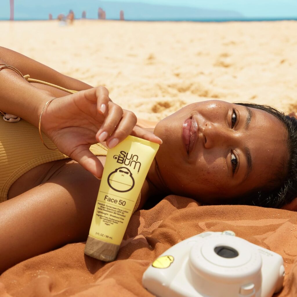 A woman with glowing skin relaxing on the beach, holding a yellow tube of Sun Bum Face 50 sunscreen. She's lying on an orange towel with a small white instant camera beside her, with the sandy shore and ocean visible in the background.